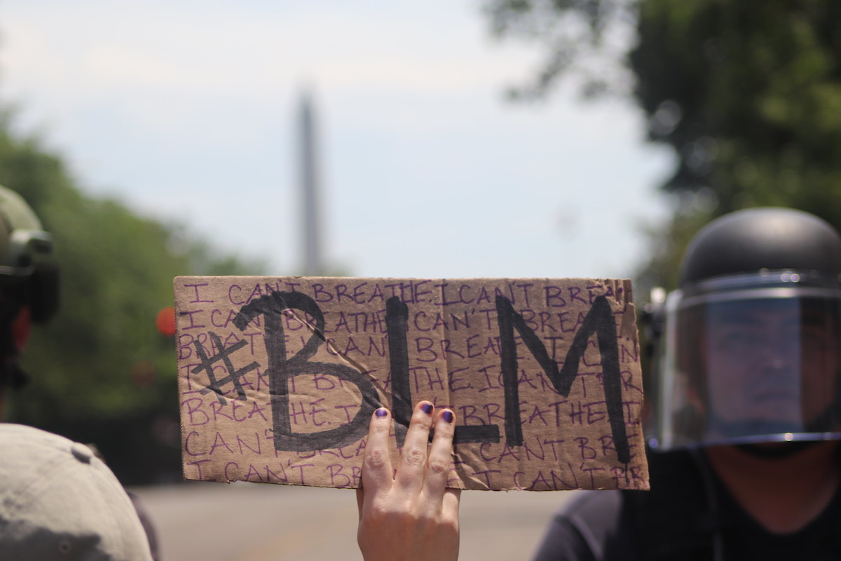 A hand holding a cardboard sign which has "I CAN'T BREATHE" repeating in purple lettering and "BLM" overtop in a bold font. Out-of-focus behind the protester are two cops in riot gear.
