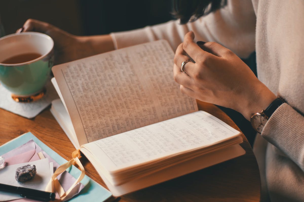 A woman has a notebook filled with writing open on a table. She holds a mug of tea in one hand.