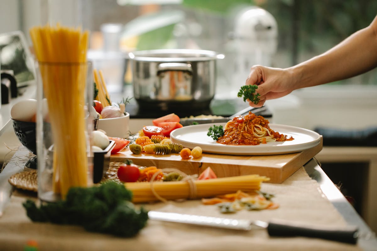 A person garnishing a plate of pasta with tomato sauce and parsley, sitting on a counter with fresh ingredients.