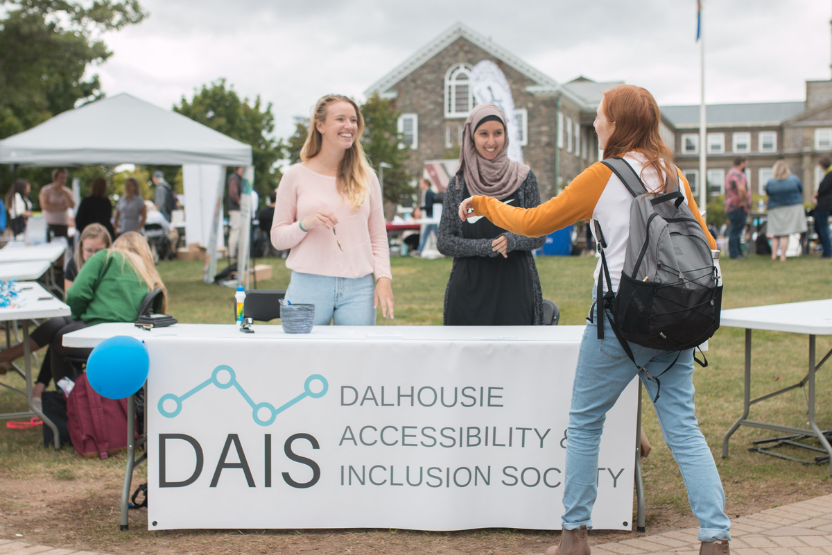 Two smiling women stand behind a table for the "Dalhousie Accessibility and Inclusion Society". They are talking to a woman on the other side of the tabel.