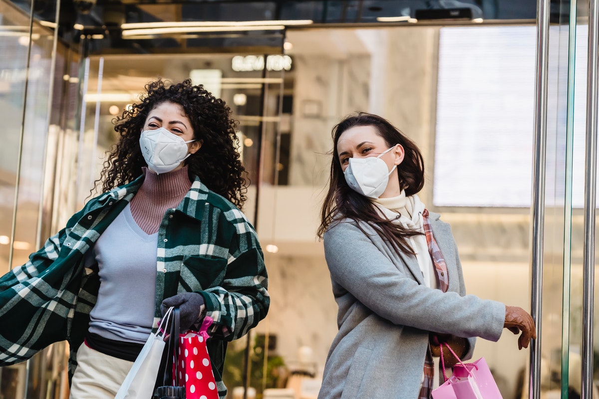 Two women wearing face masks and holding shopping bags exit a store.