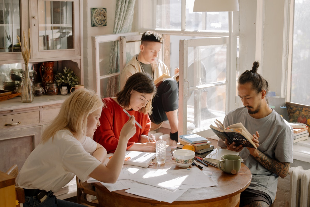 A group of young adults sit around a table in a sunny kitchen. One is painting, one is journalling, and two are reading.
