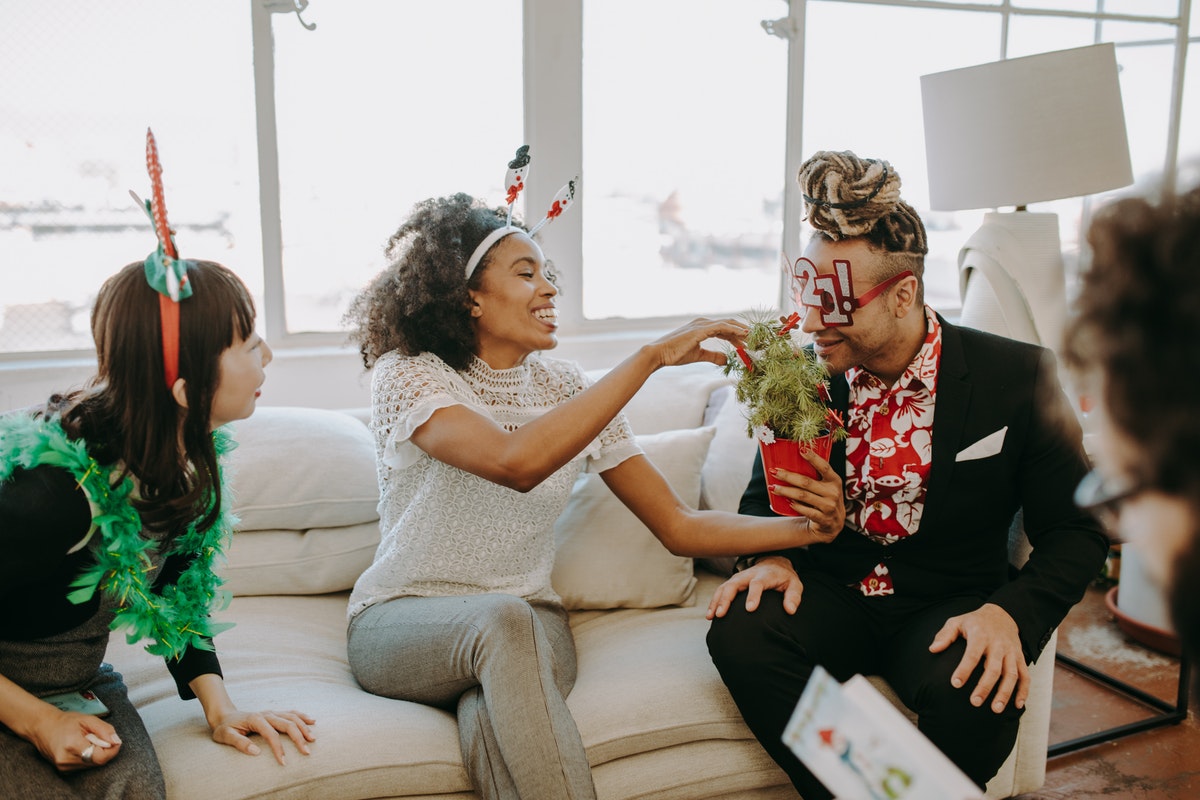 Four adults wearing Christmas- and New Years -themed accessories sit around a living room. They are playing some sort of game involving sniffing a plant.