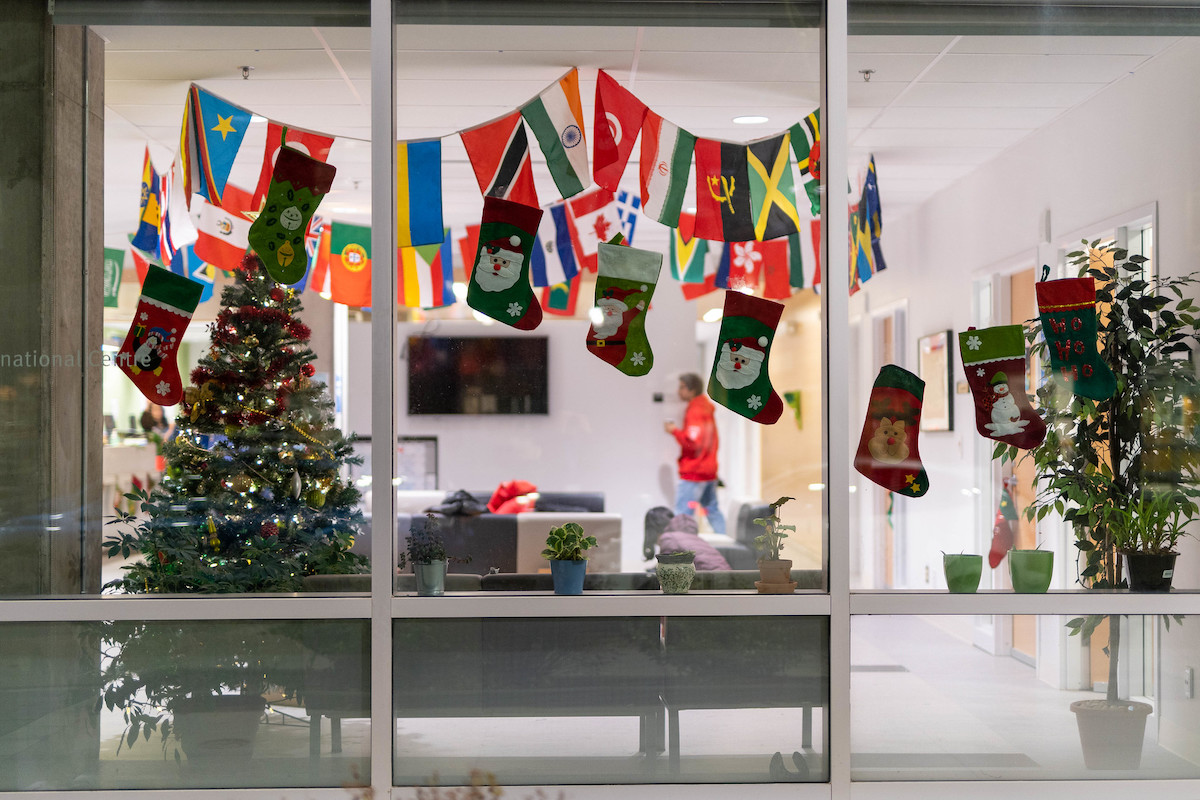 A look through the windows of the International Centre, where stockings with Santa on it hang in front of a banner of international flags, and a Christmas tree stands in the corner.