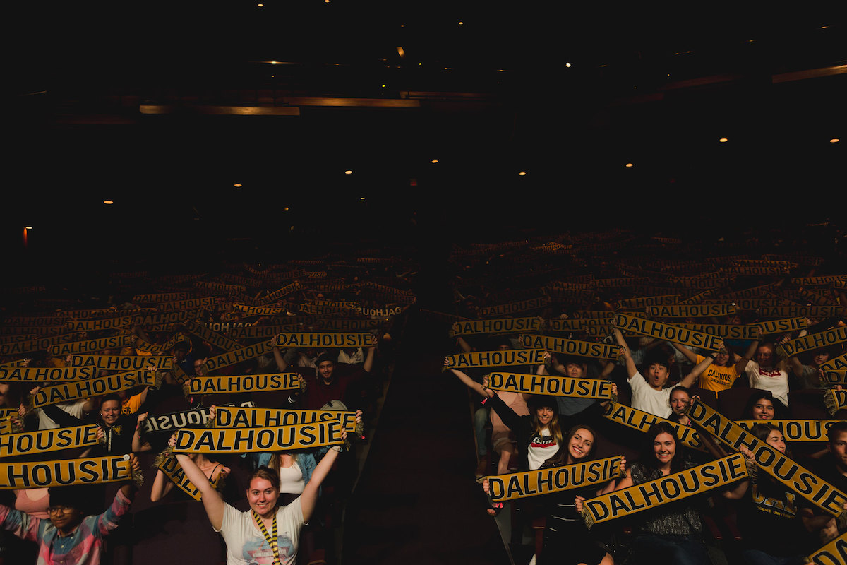 A group of Dalhousie students are seated in an auditorium, holding up scarves that read "DALHOUSIE" and smiling at the camera.