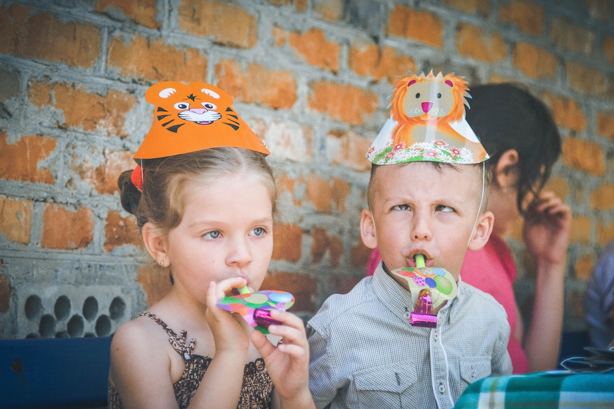 Two children sitting beside each other with party hats on their heads and party blowers in their mouths
