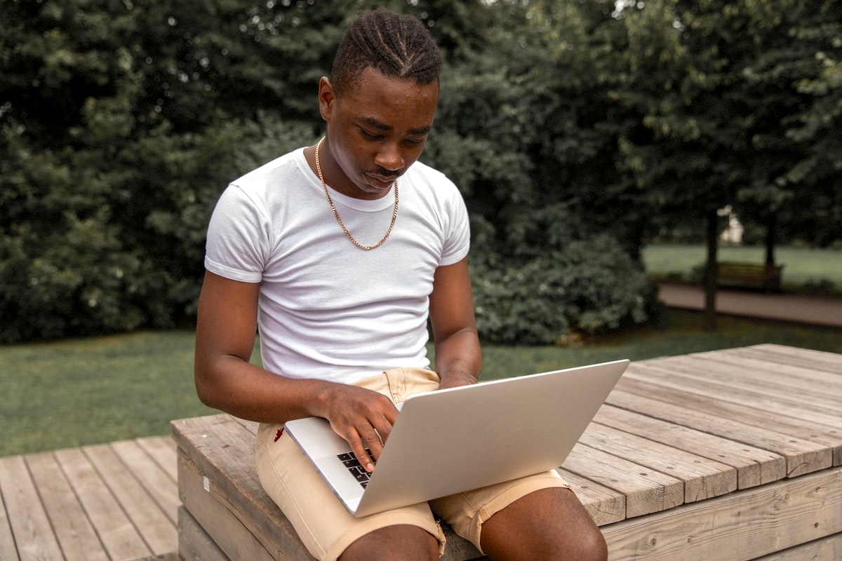 A Black man in a white t-shirt and shorts sits outside on a bench with a laptop on his lap.
