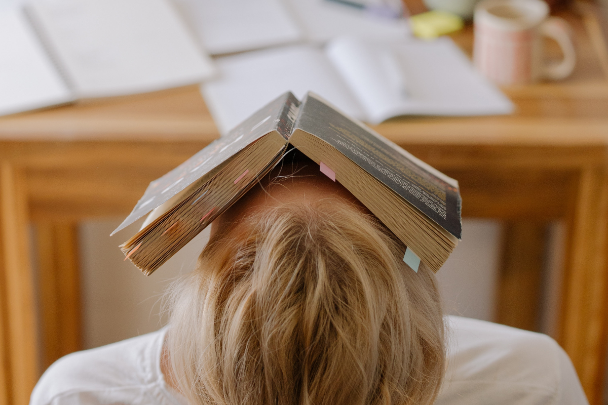 A blonde woman in a white T-shirt sits in front of a wooden table covered with school supplies. Her head is leaned back, and a book is laying open on top of her face.