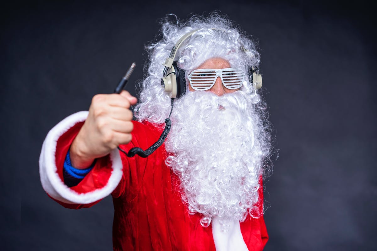 Person dressed in a Santa Claus costume with white curly hair and beard, crazy sunglasses, and wearing headphones