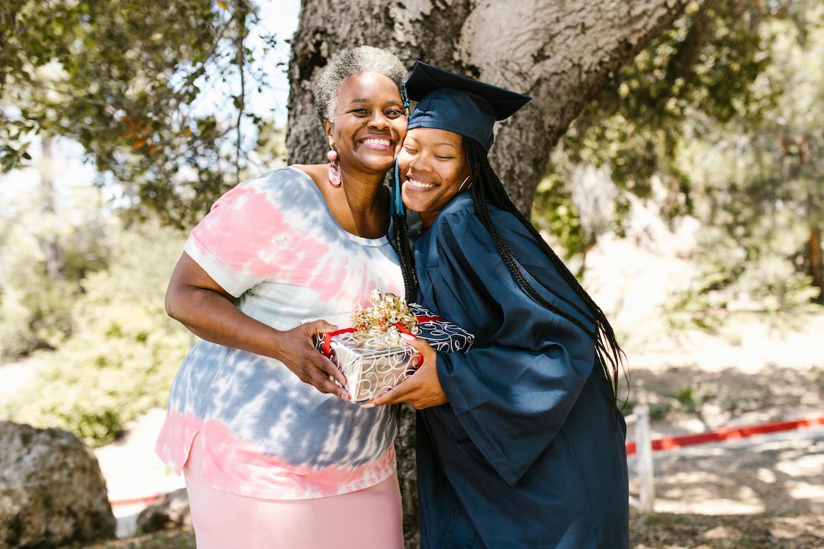 A young woman in graduation robes hugging an older woman with both smiling while holding a gift
