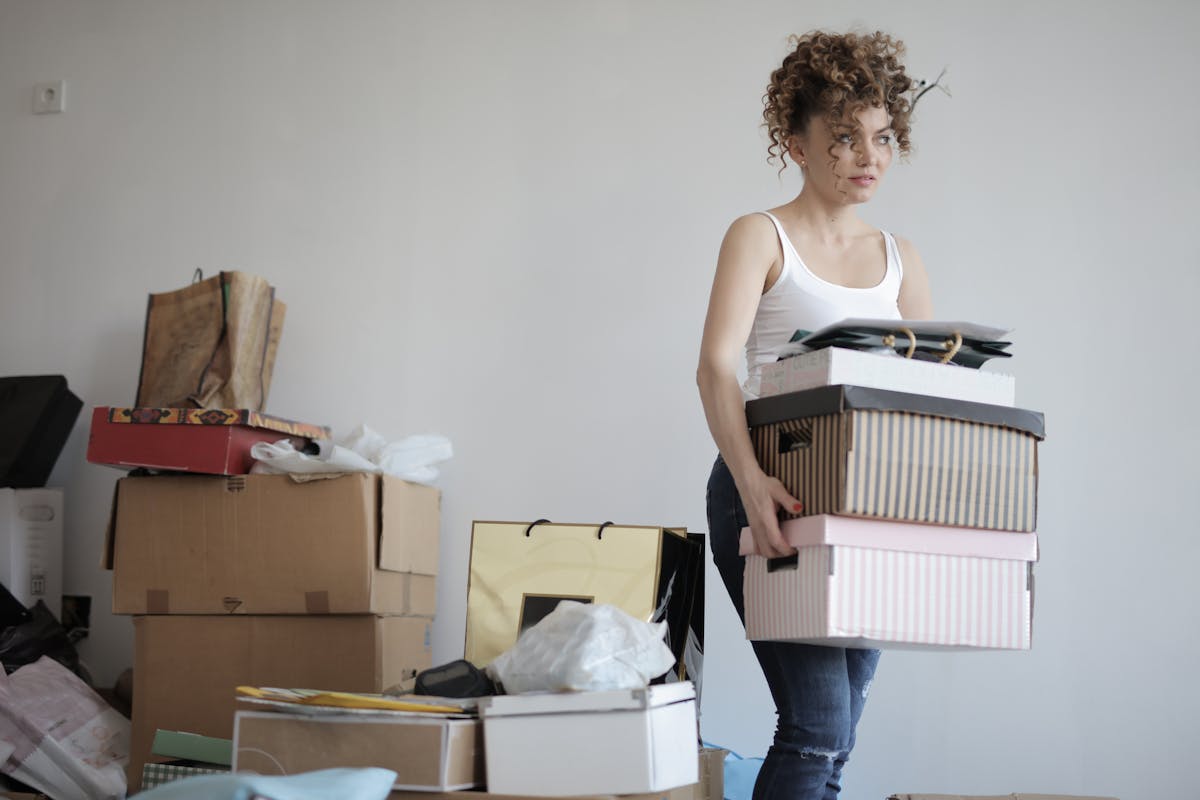 A woman with curly hair holding a stack of boxes with more boxes piled up behind her