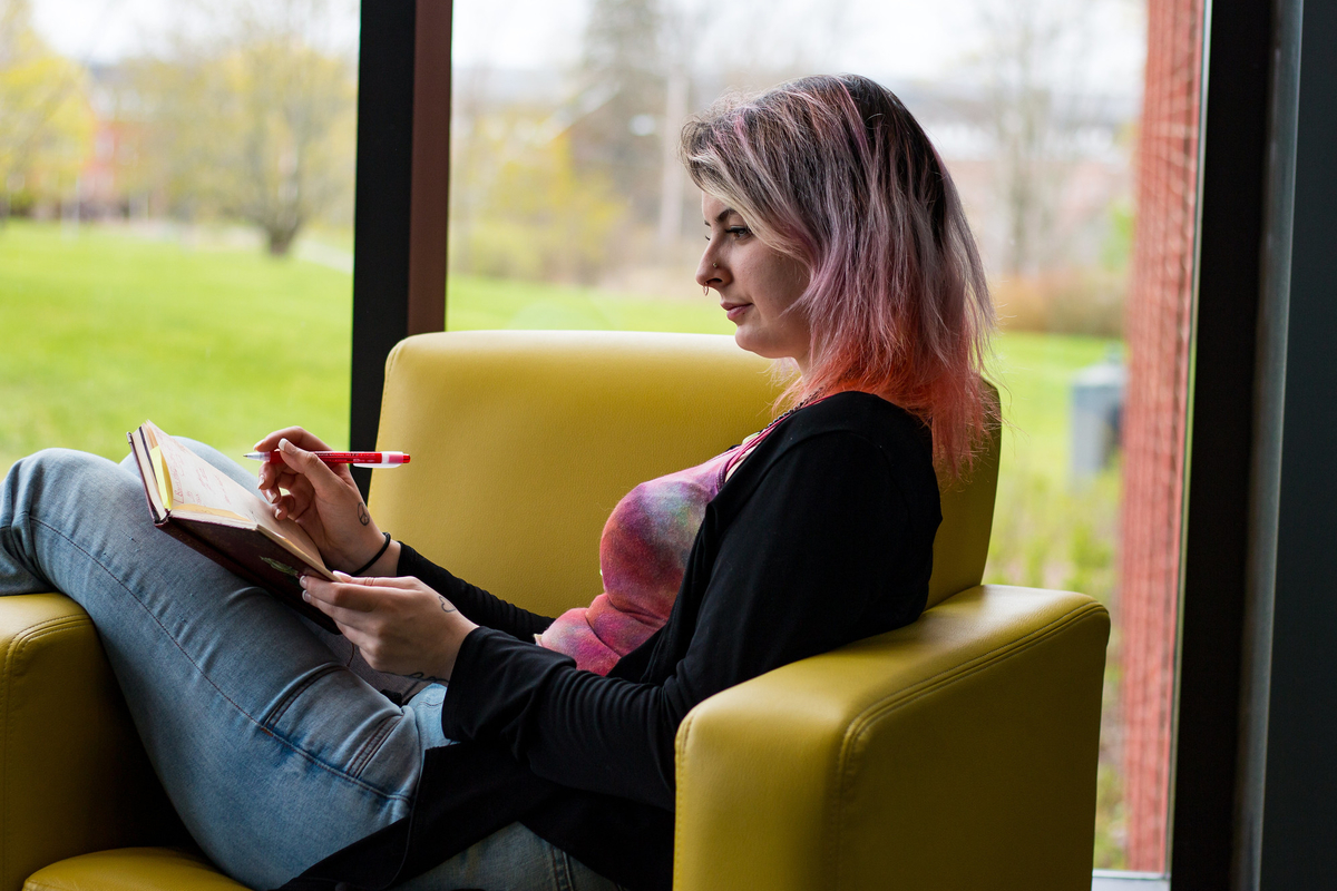 A young woman sits in a yellow leather chair with a notebook and pen in her lap.