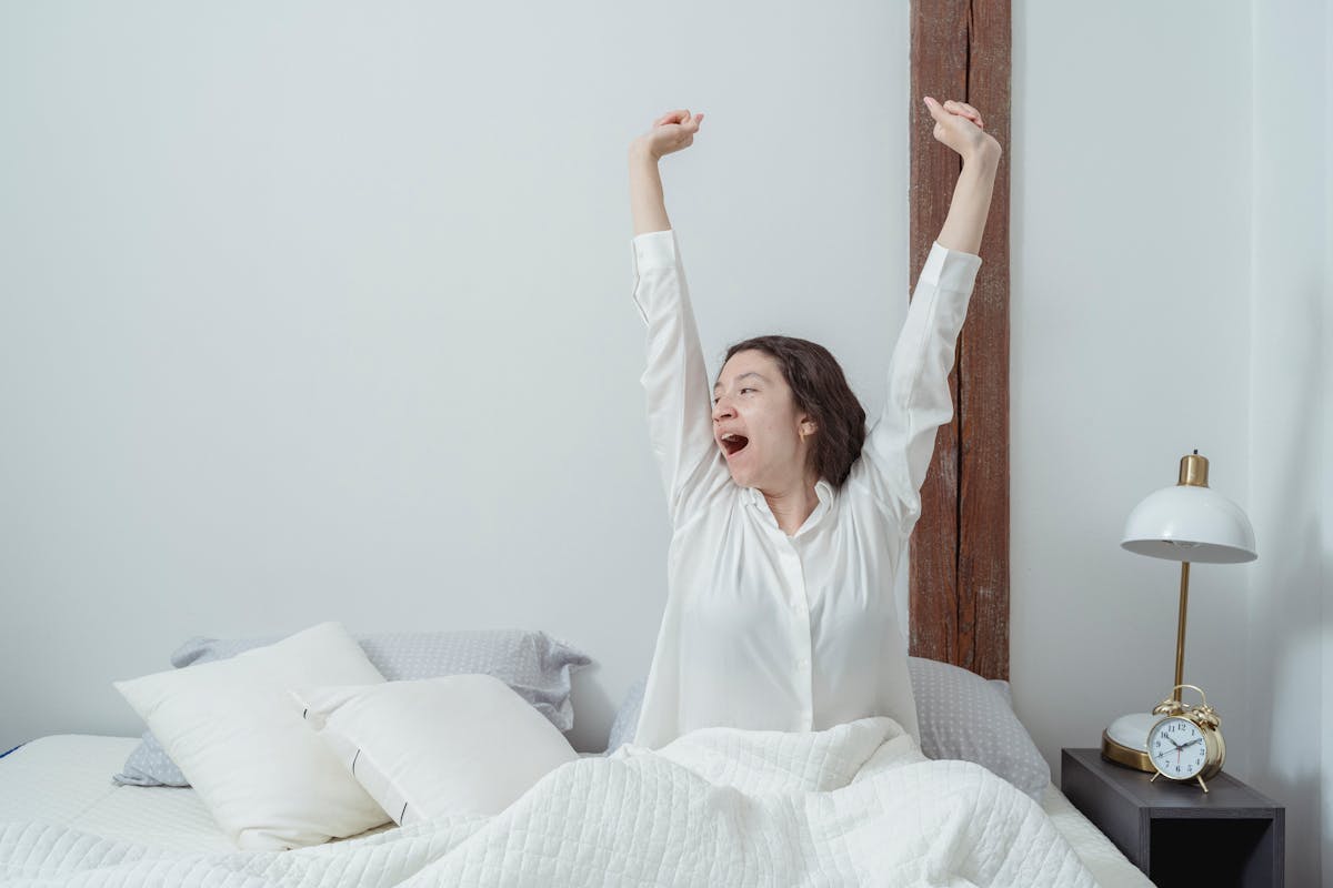 A young woman sitting up in a white bed, stretching and yawning