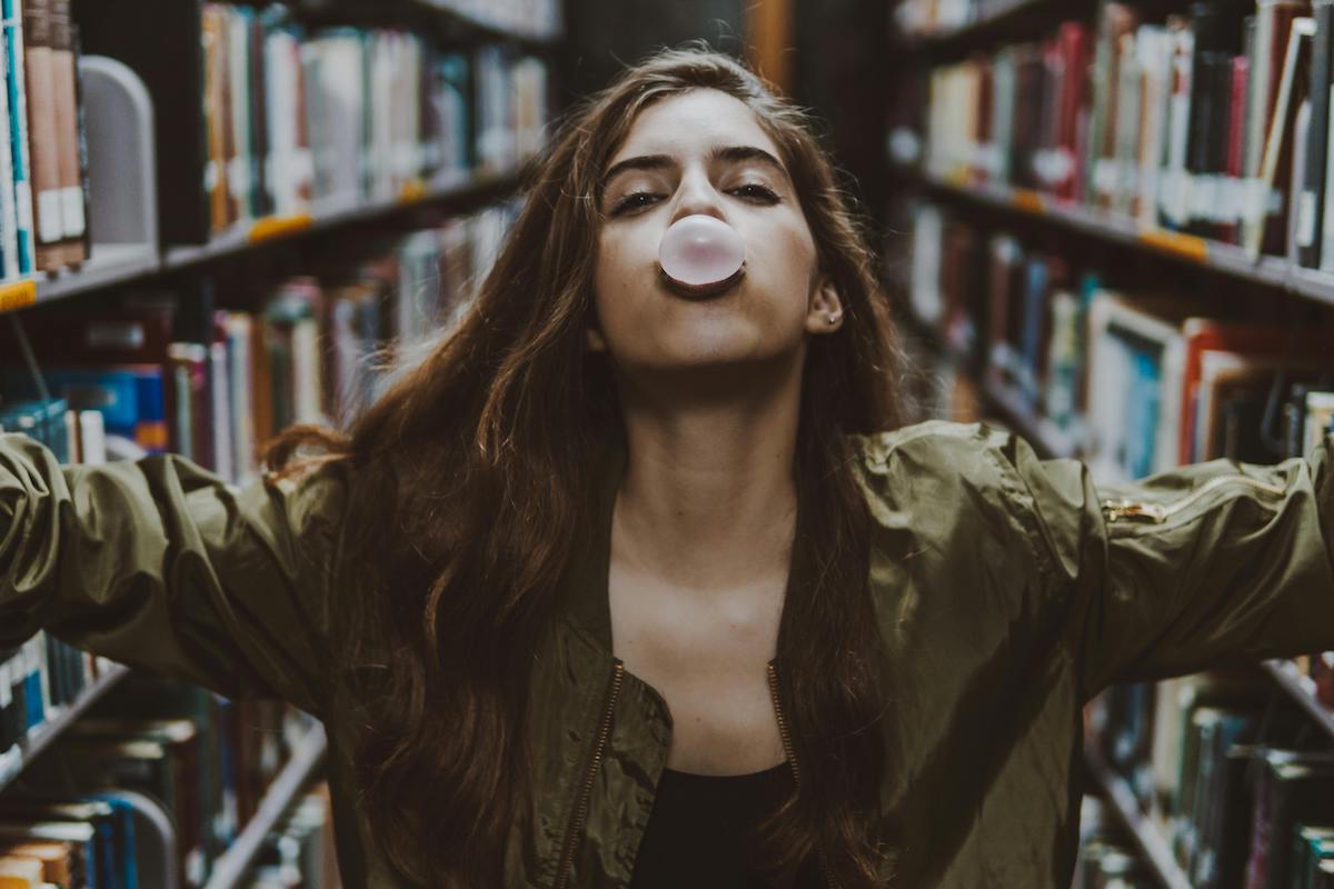 A young woman in a green jacket blowing a chewing gum bubble, standing between shelves of books