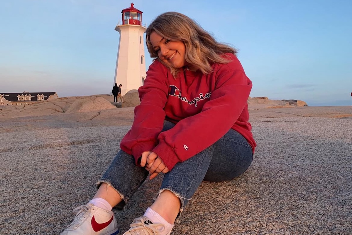 Young woman in a red sweatshirt and jeans sitting on flat rocks in front of a lighthouse