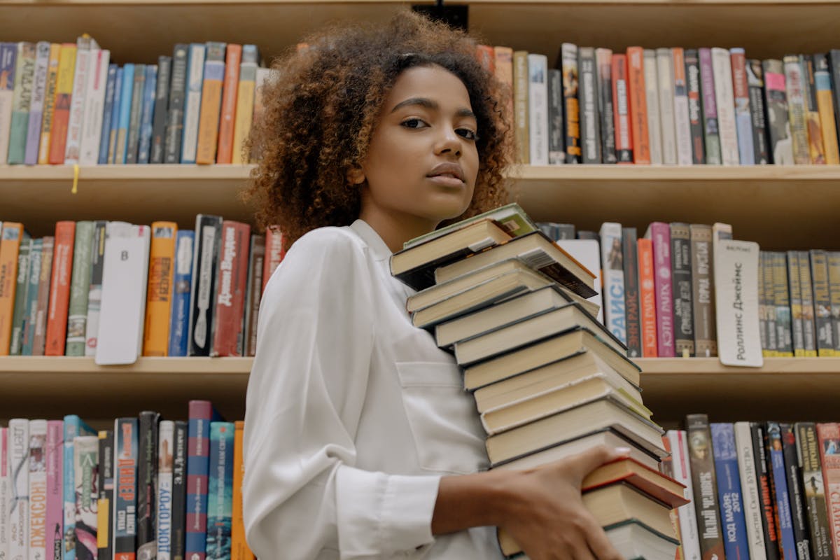 A young woman with dark curly hair, wearing a white shirt, holds a stack of books in front of her while standing in front of shelves full of books.