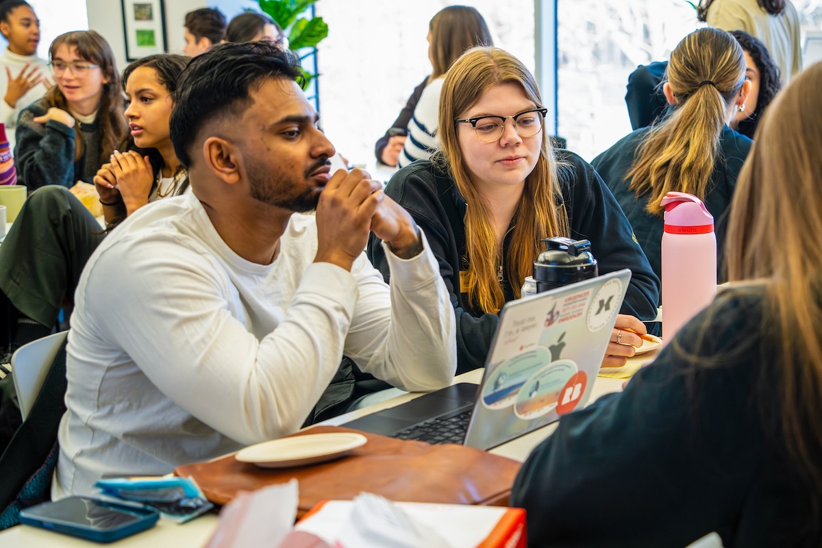 A group of students sitting at a table looking at a laptop with other students talking in the background.
